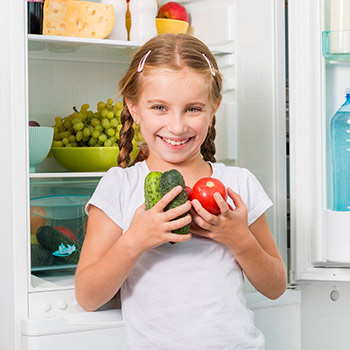 Lächelndes Mädchen hält Gemüse vor geöffnetem Kühlschrank